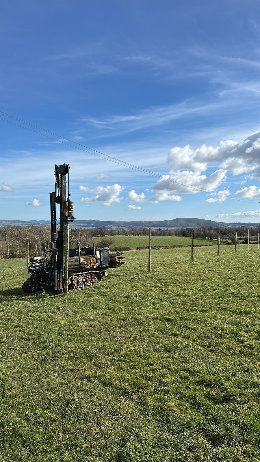 A tracked post knocker driving stakes for a new fence line across open pasture with hills beyond