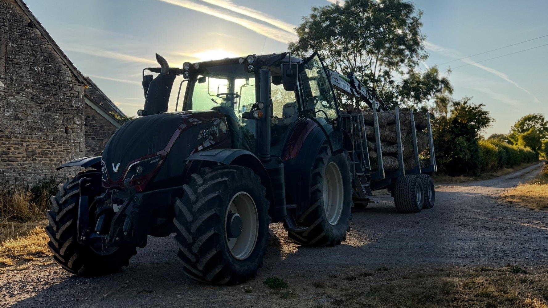 A JRF Contracting Valtra tractor with a Botex timber crane and a loaded log trailer standing on a farm track beside a stone barn at sunset