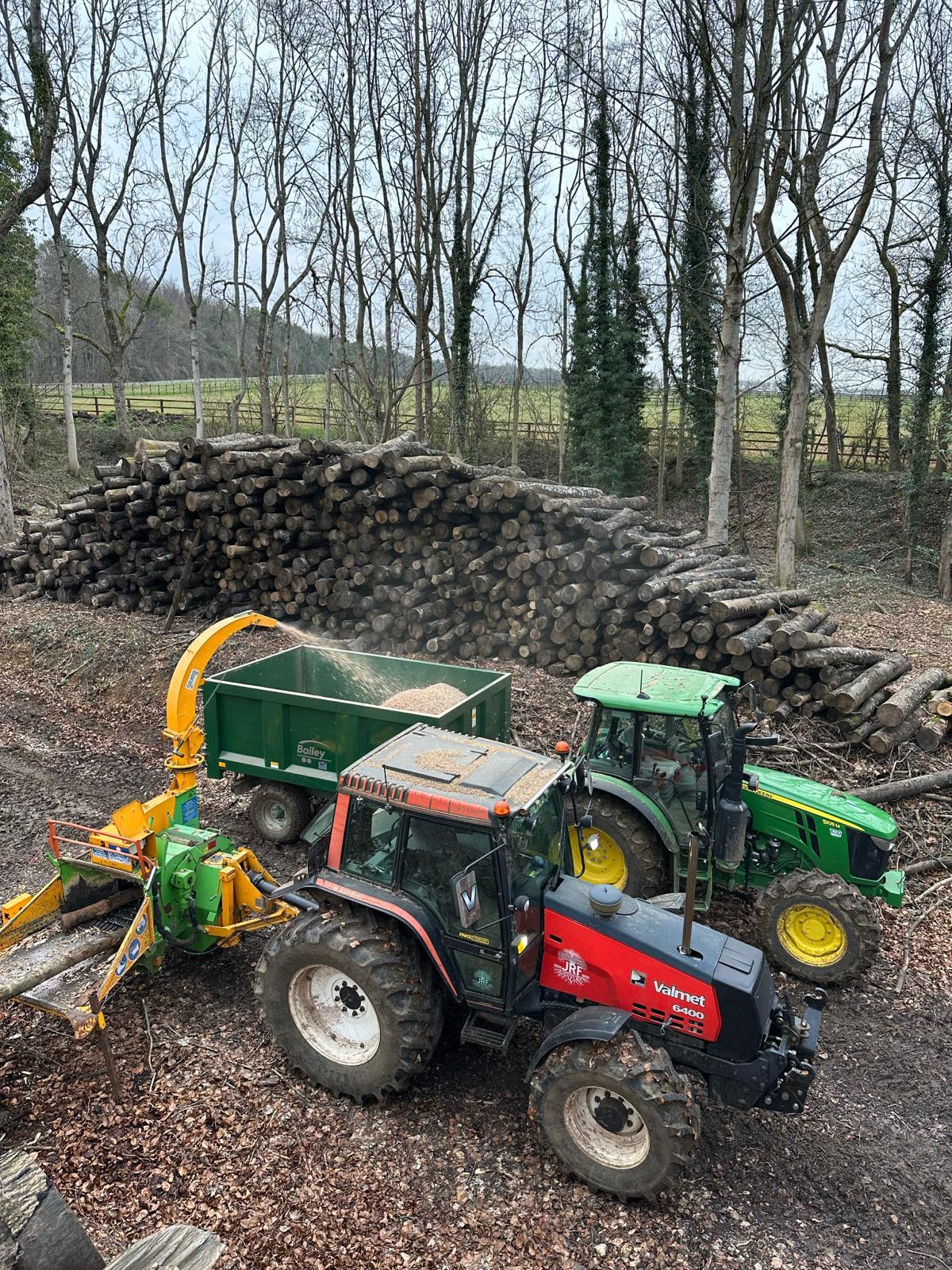 Two tractors and a PTO chipper filling a trailer beside a long stack of cordwood in woodland
