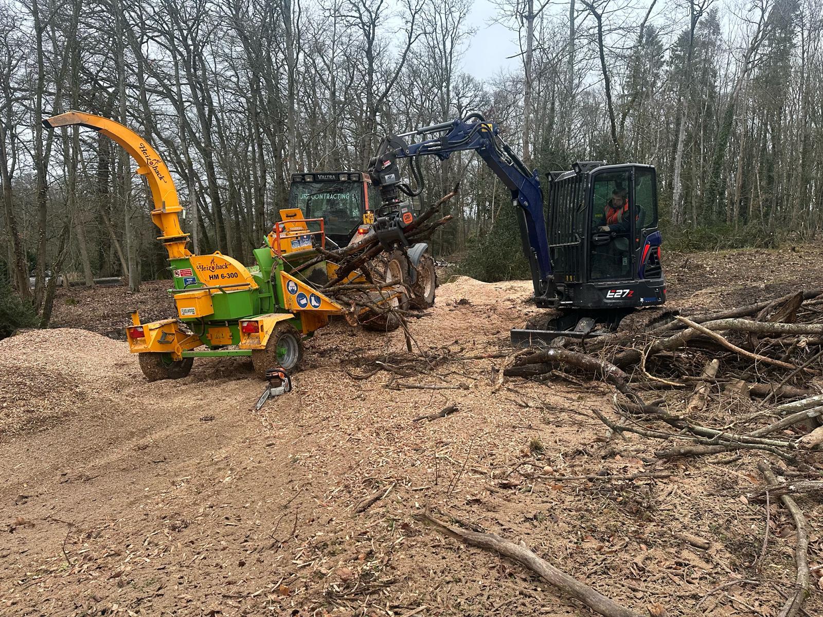 A wood chipper and a tracked excavator working together on a cleared woodland floor covered in fresh chip