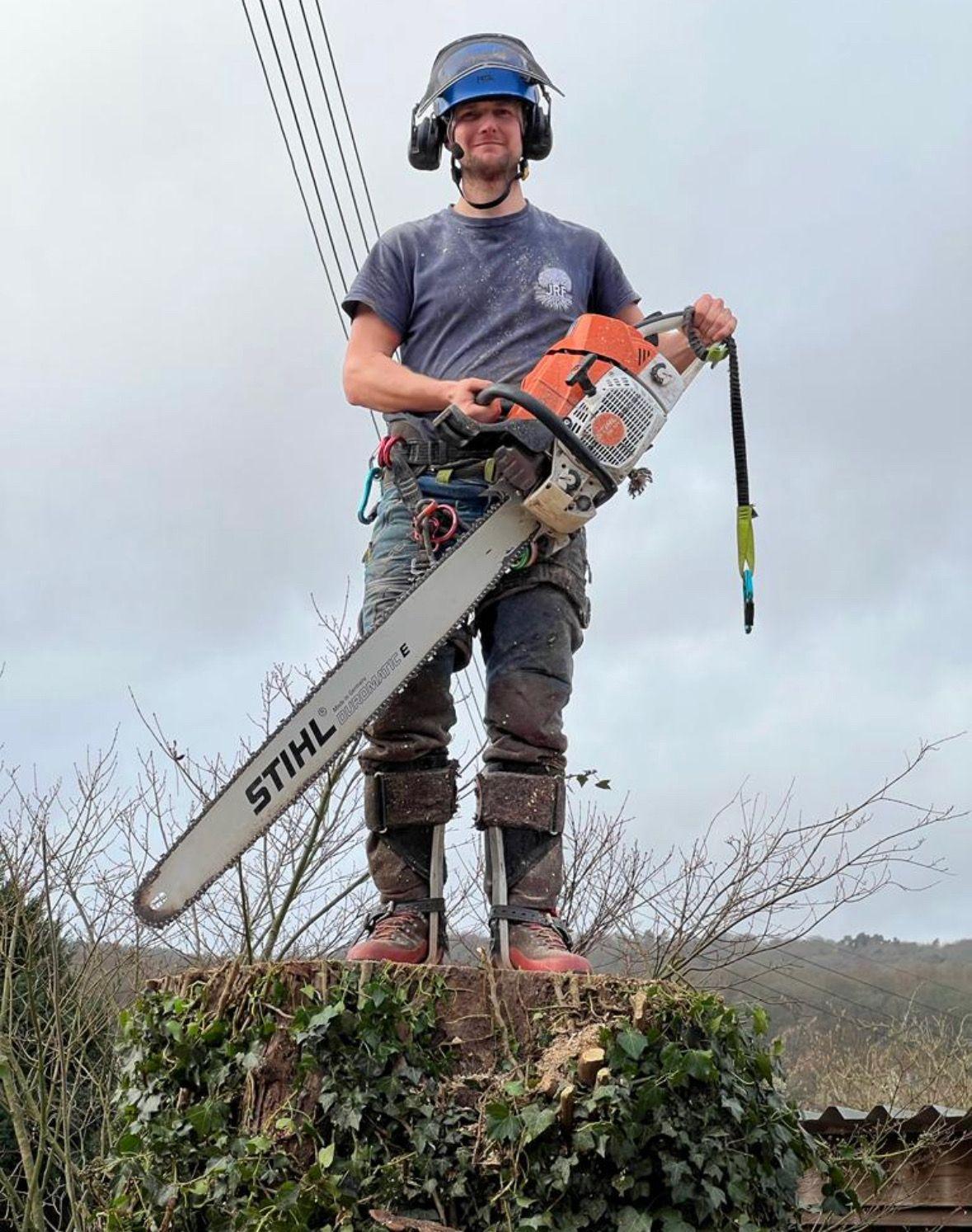 Joe Finnemore in full chainsaw protective equipment — helmet with visor and ear defenders, chainsaw trousers, chainsaw boots and harness — holding a chainsaw on a felled stump